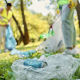 Two people wearing gloves and safety vests pick up litter in a park. In the foreground, a crumpled can and plastic bag are visible on the grass, whilst one person collects rubbish in a clear bag.