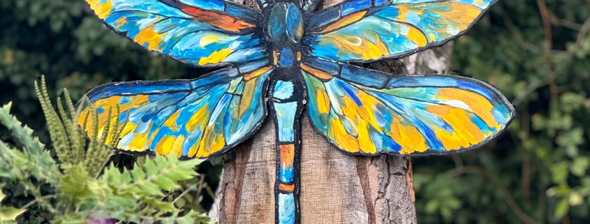 A colorful, large dragonfly art piece with blue and yellow wings is mounted on a vertical tree stump in a garden. Purple flowers are blurred in the foreground and green foliage is visible in the background.