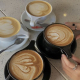 Four cups of latte with heart-shaped latte art are on a speckled table. Two cups are white, two are black. A person with rings on their fingers is holding one of the black cups. Spoons rest on the saucers.