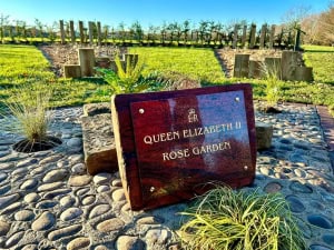 A polished plaque reading "Queen Elizabeth II Rose Garden" stands on a stone path, with green grass, small plants, and young trees in a sunny garden behind it.